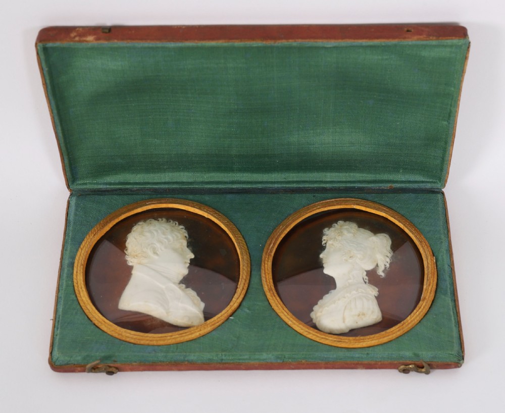A pair of white wax profile portrait reliefs of a man and a woman framed in circular brass bezels and displayed inside a green fabric-lined presentation case.