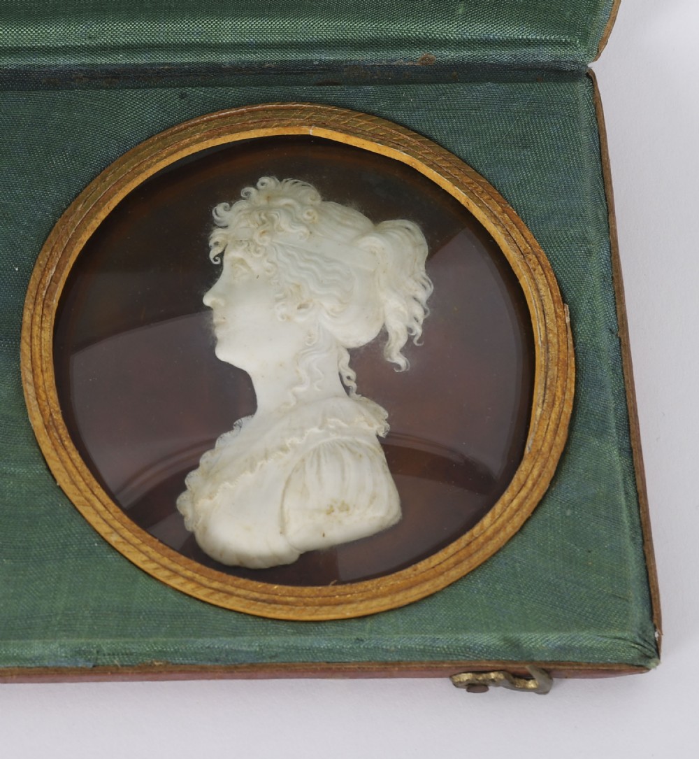 A pair of white wax profile portrait reliefs of a man and a woman framed in circular brass bezels and displayed inside a green fabric-lined presentation case. View III