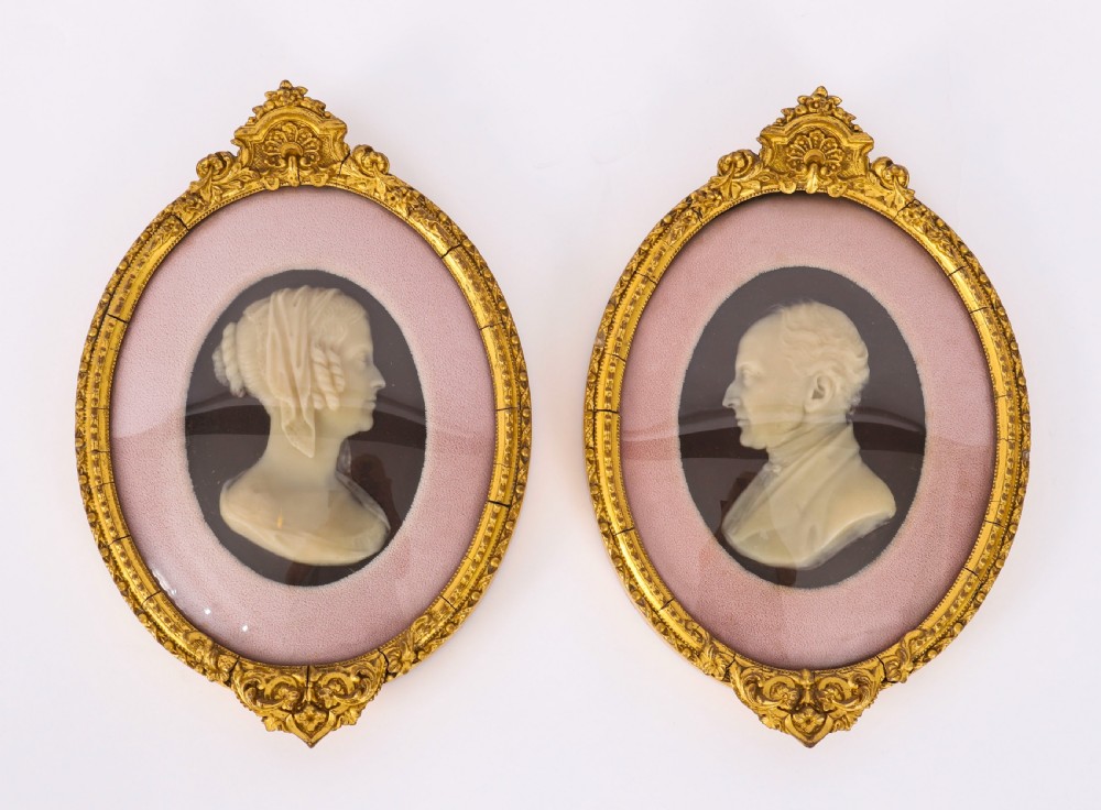 A pair of oval gilt-framed 19th-century wax portrait reliefs featuring male and female profile busts set against dark backgrounds with pink fabric mounts.
