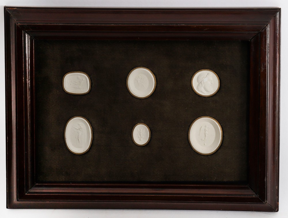 A dark wooden moulding frame housing six white plaster classical intaglio impressions set against a dark brown velvet background.