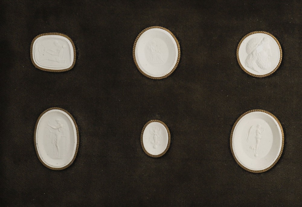 A dark wooden moulding frame housing six white plaster classical intaglio impressions set against a dark brown velvet background. View II