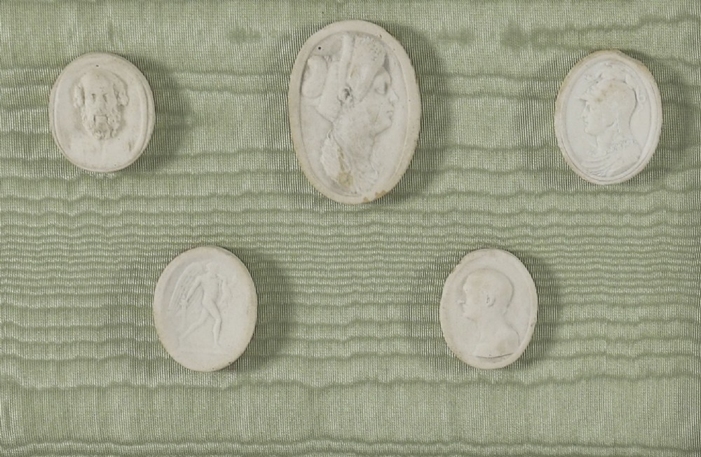 A pair of framed displays containing white plaster intaglio relief medallions mounted on green patterned fabric inside gilt frames. View VI
