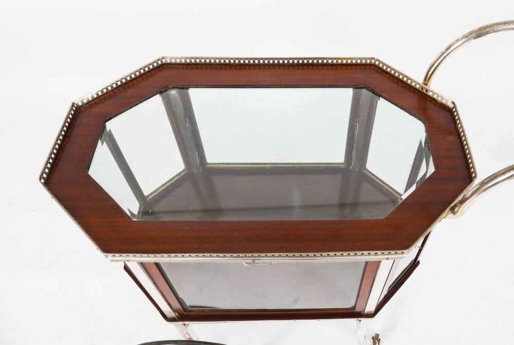 An Art Deco drinks trolley featuring an octagonal glass top with metal gallery trim, wooden side frames, spoked wheels, and a drop-down door. View X