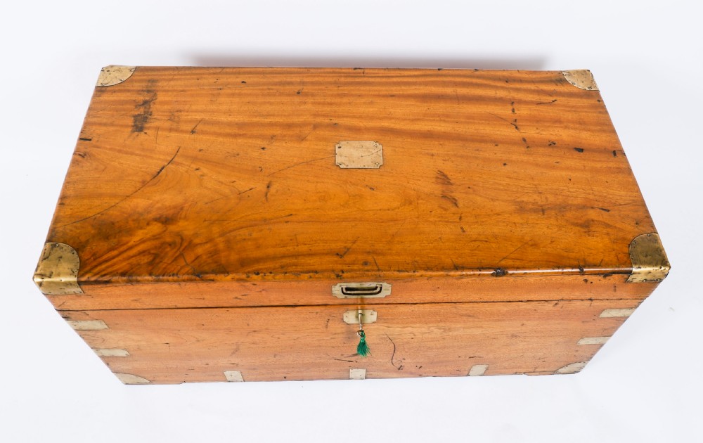 A rectangular 19th-century warm brown camphorwood military campaign trunk featuring flush brass corner brackets, protective metal straps, side drop handles, and a central brass keyhole escutcheon. View V