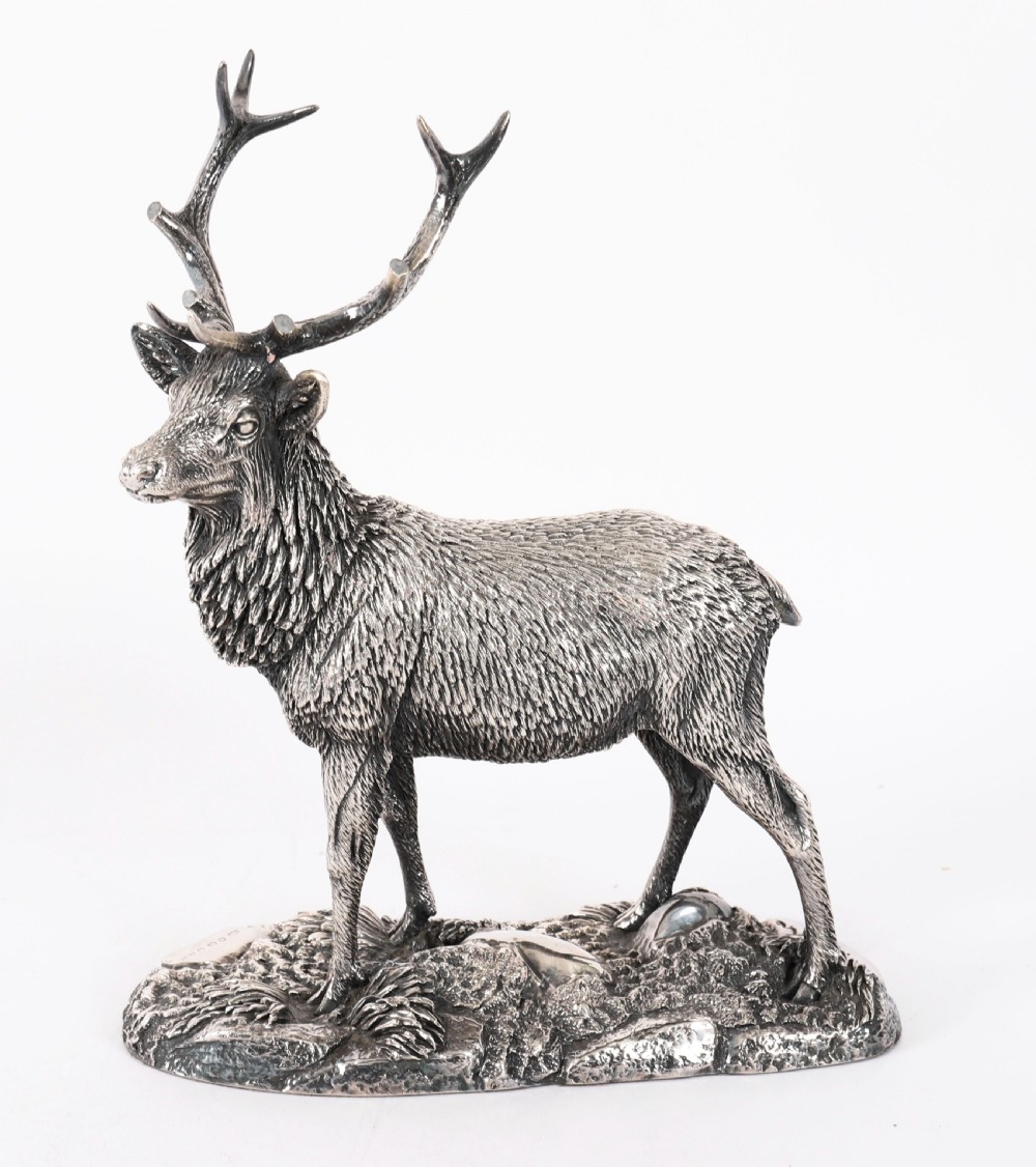A detailed sterling silver sculpture of a standing stag with branching antlers positioned on a textured rocky ground base.