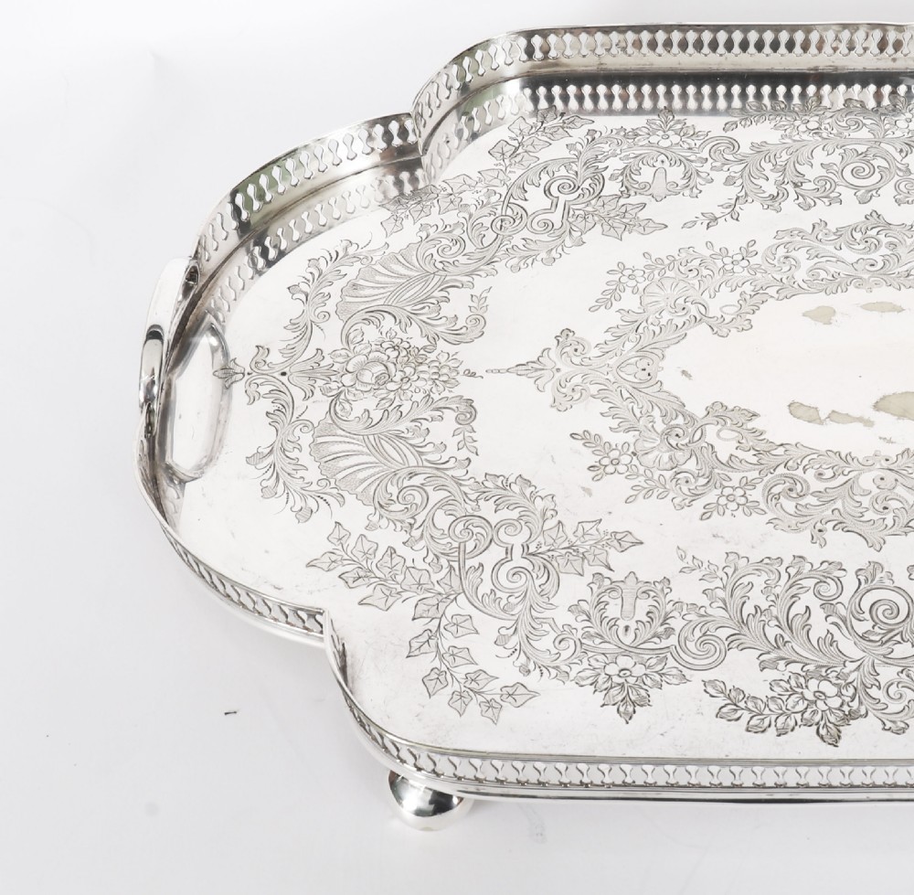 An Edwardian silver-plated gallery tray featuring a pierced rim, engraved foliate scrollwork, twin handles, and ball feet. View IV