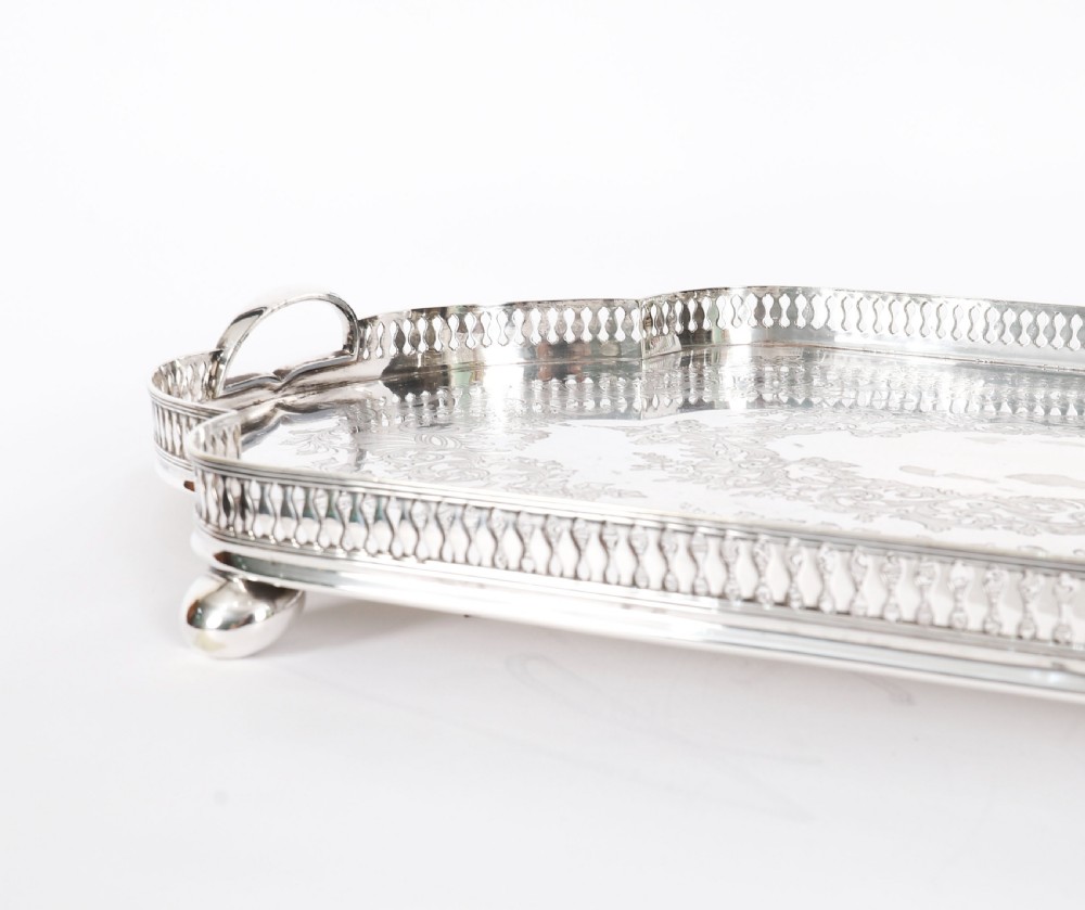 An Edwardian silver-plated gallery tray featuring a pierced rim, engraved foliate scrollwork, twin handles, and ball feet. View IX