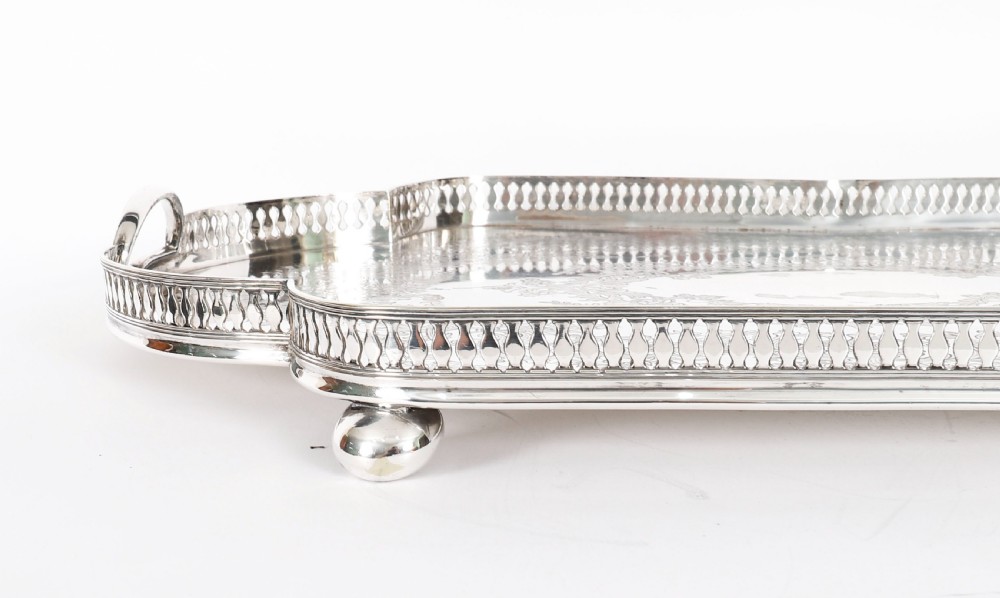 An Edwardian silver-plated gallery tray featuring a pierced rim, engraved foliate scrollwork, twin handles, and ball feet. View XII