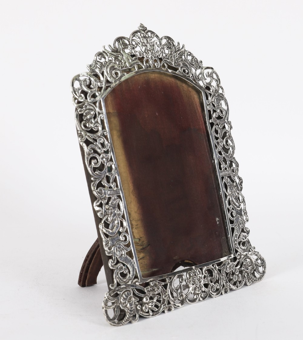 An antique sterling silver photo frame featuring openwork scrollwork detailing and a dark wooden easel back.