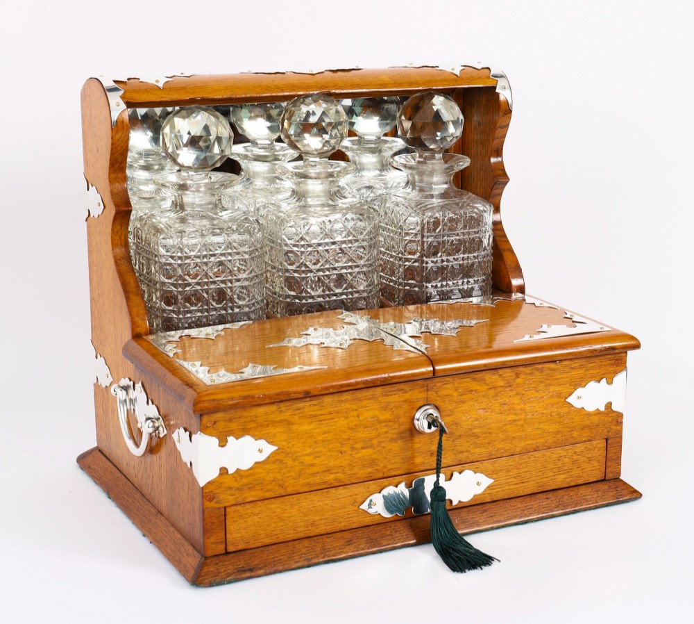 An antique blonde oak tantalus featuring three square cut-glass decanters set against a mirror back, decorative silver-plated corner mounts, and a lower drawer with a key.