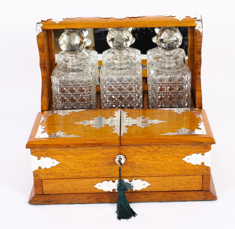 An antique blonde oak tantalus featuring three square cut-glass decanters set against a mirror back, decorative silver-plated corner mounts, and a lower drawer with a key. View II