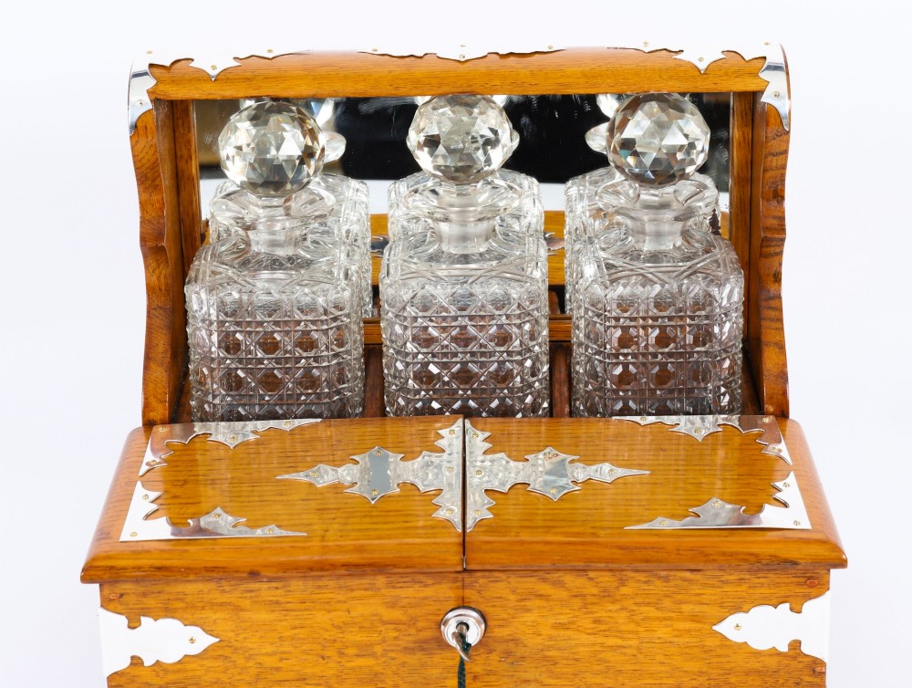 An antique blonde oak tantalus featuring three square cut-glass decanters set against a mirror back, decorative silver-plated corner mounts, and a lower drawer with a key. View III
