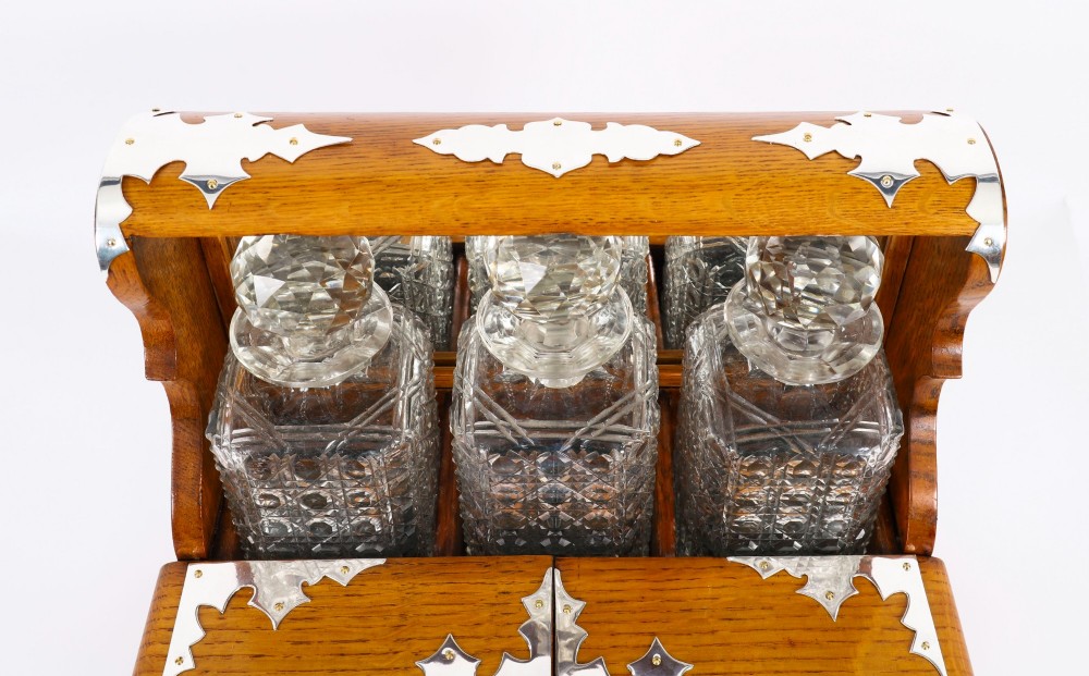 An antique blonde oak tantalus featuring three square cut-glass decanters set against a mirror back, decorative silver-plated corner mounts, and a lower drawer with a key. View IV