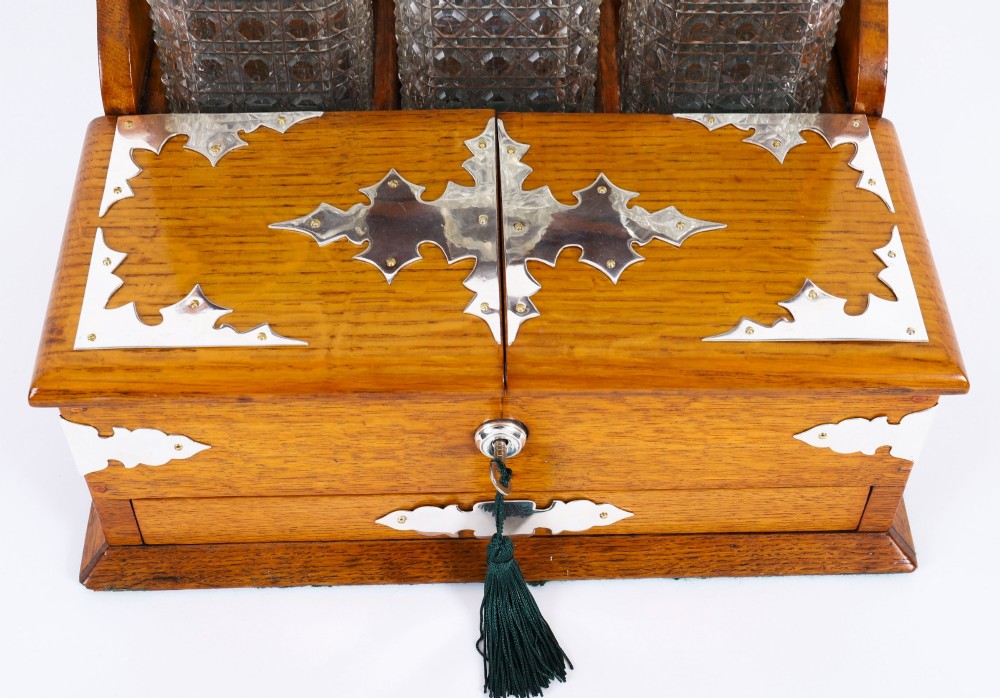 An antique blonde oak tantalus featuring three square cut-glass decanters set against a mirror back, decorative silver-plated corner mounts, and a lower drawer with a key. View V