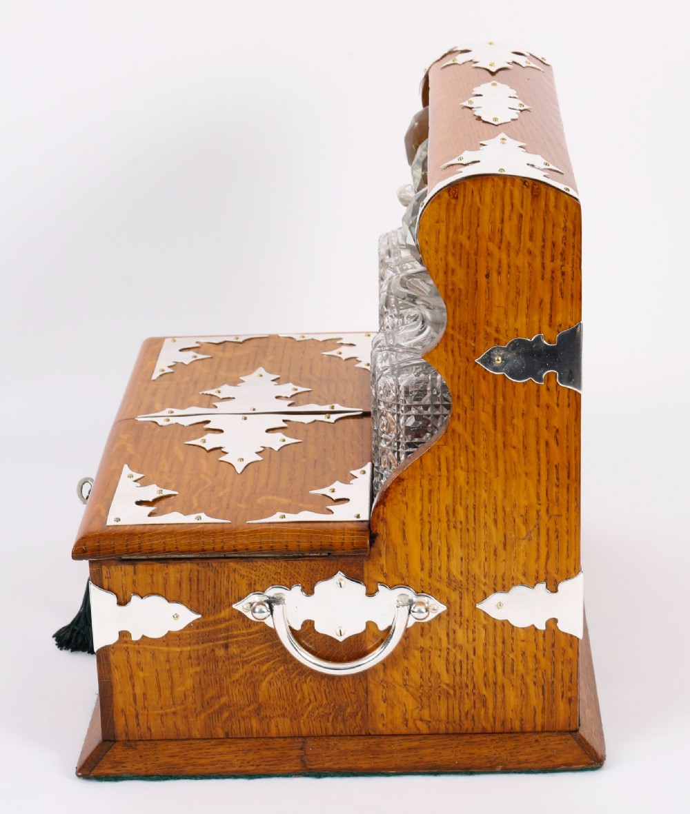 An antique blonde oak tantalus featuring three square cut-glass decanters set against a mirror back, decorative silver-plated corner mounts, and a lower drawer with a key. View VI