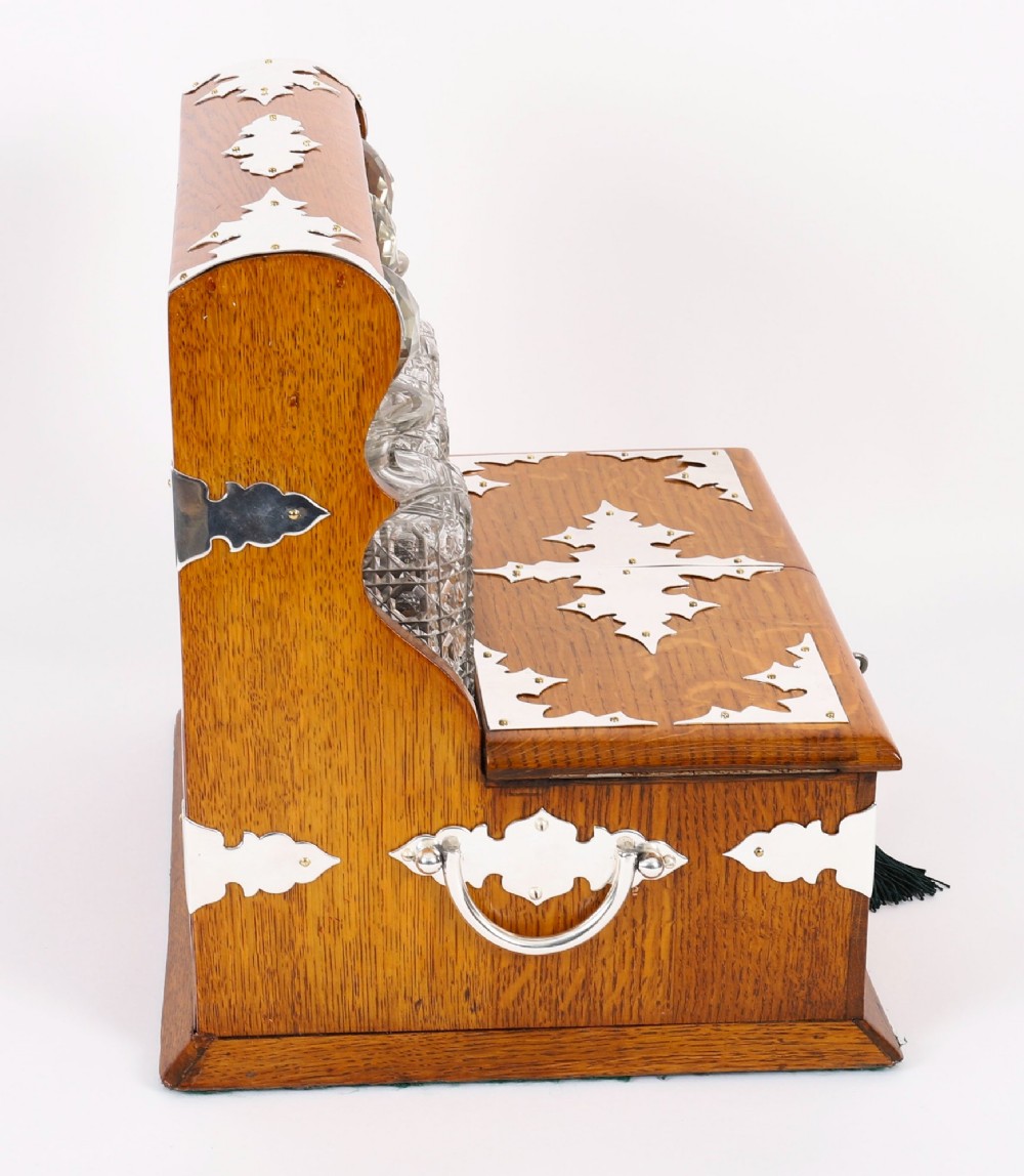 An antique blonde oak tantalus featuring three square cut-glass decanters set against a mirror back, decorative silver-plated corner mounts, and a lower drawer with a key. View VII