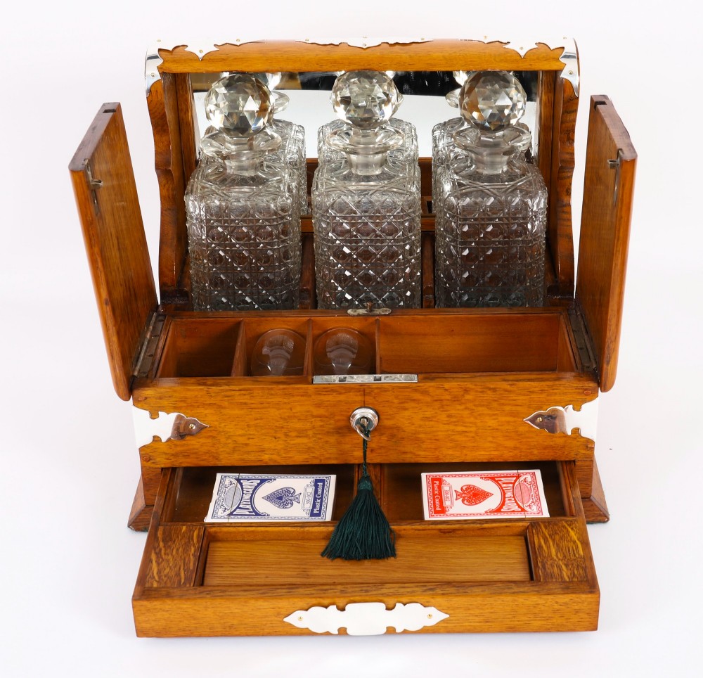 An antique blonde oak tantalus featuring three square cut-glass decanters set against a mirror back, decorative silver-plated corner mounts, and a lower drawer with a key. View VIII