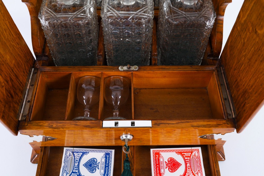 An antique blonde oak tantalus featuring three square cut-glass decanters set against a mirror back, decorative silver-plated corner mounts, and a lower drawer with a key. View X