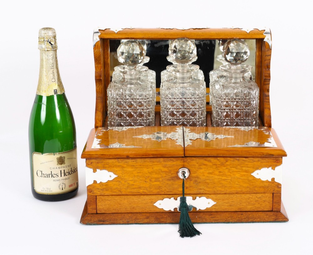 An antique blonde oak tantalus featuring three square cut-glass decanters set against a mirror back, decorative silver-plated corner mounts, and a lower drawer with a key. View XV