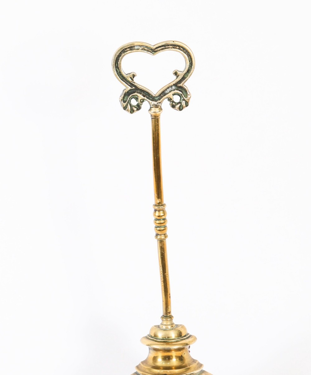 A 19th-century Victorian brass door stop featuring a tall shaft topped with a heart-shaped handle and supported by a heavy, tiered circular base. View VII