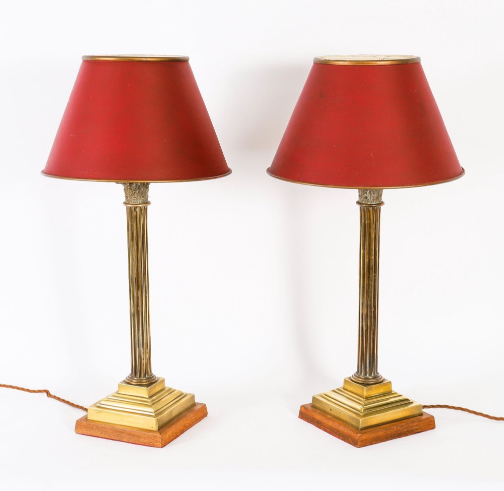 A pair of late nineteenth-century brass Corinthian column table lamps featuring fluted stems, stepped bases, wooden plinths, and conical red lampshades.