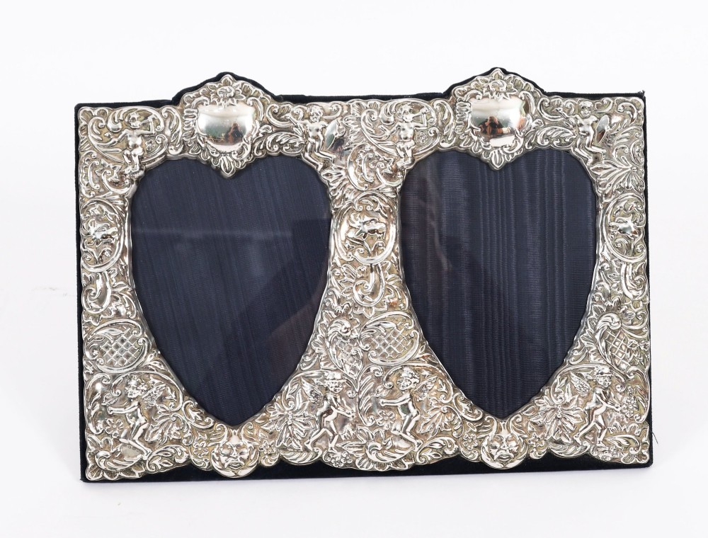 An antique sterling silver double aperture photo frame featuring heart-shaped openings, intricate repousse floral scrollwork, and cherub details mounted on dark velvet.