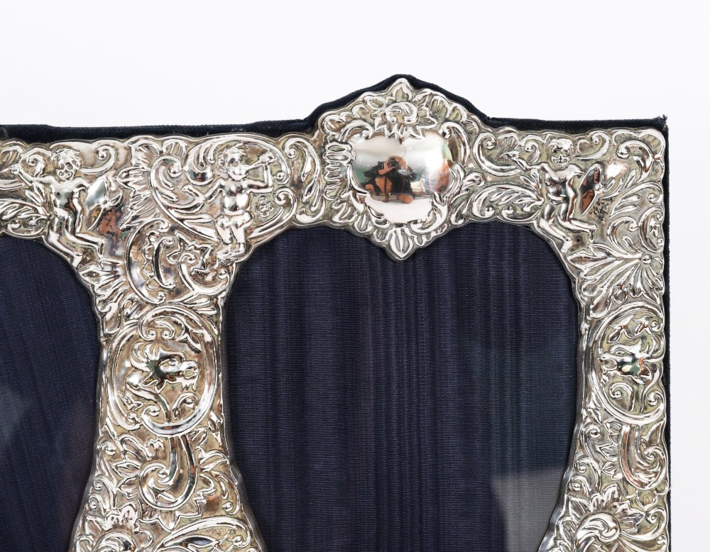 An antique sterling silver double aperture photo frame featuring heart-shaped openings, intricate repousse floral scrollwork, and cherub details mounted on dark velvet. View III