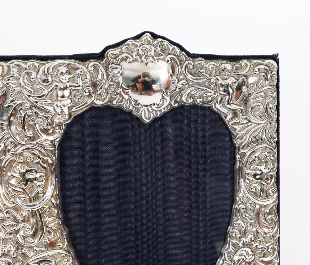 An antique sterling silver double aperture photo frame featuring heart-shaped openings, intricate repousse floral scrollwork, and cherub details mounted on dark velvet. View V