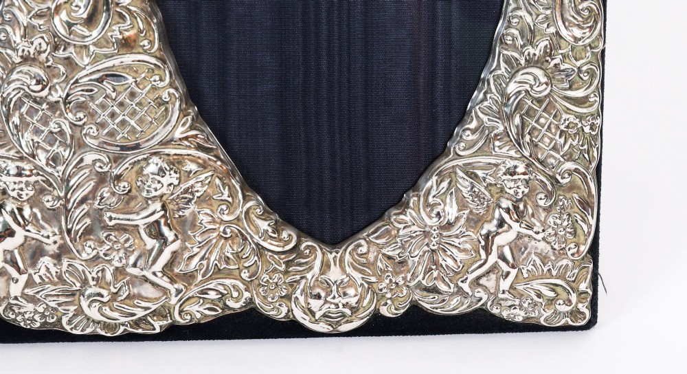 An antique sterling silver double aperture photo frame featuring heart-shaped openings, intricate repousse floral scrollwork, and cherub details mounted on dark velvet. View VIII