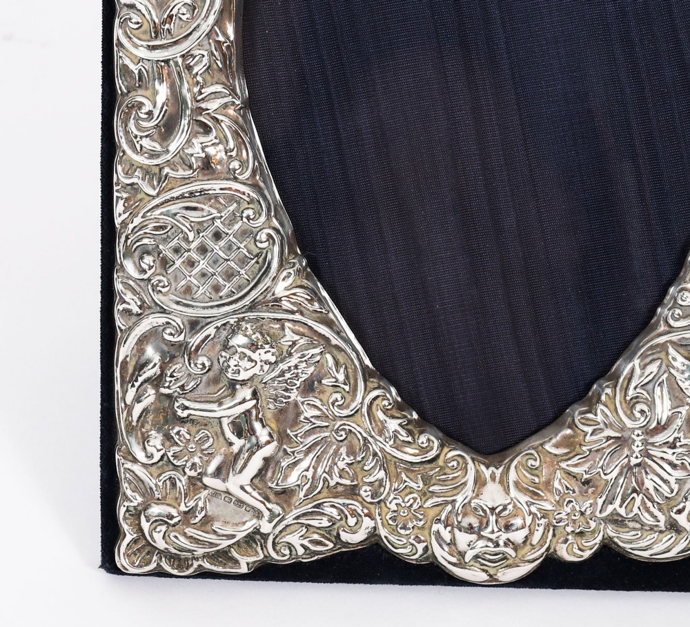 An antique sterling silver double aperture photo frame featuring heart-shaped openings, intricate repousse floral scrollwork, and cherub details mounted on dark velvet. View IX