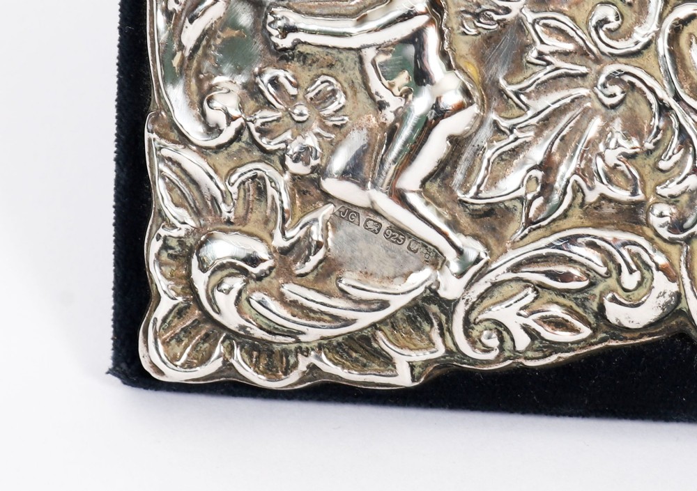 An antique sterling silver double aperture photo frame featuring heart-shaped openings, intricate repousse floral scrollwork, and cherub details mounted on dark velvet. View X