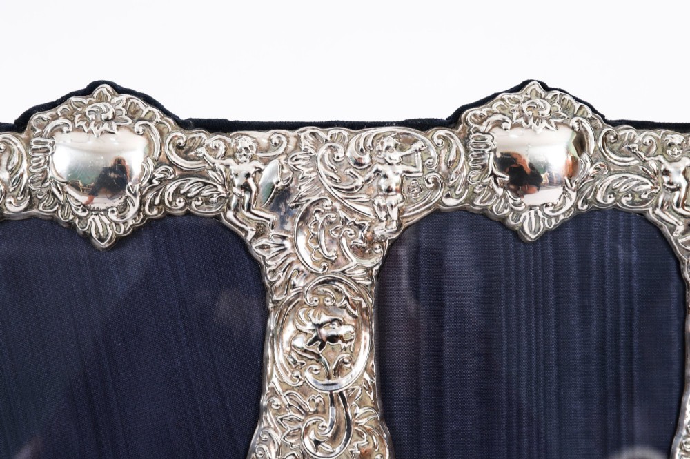 An antique sterling silver double aperture photo frame featuring heart-shaped openings, intricate repousse floral scrollwork, and cherub details mounted on dark velvet. View XI