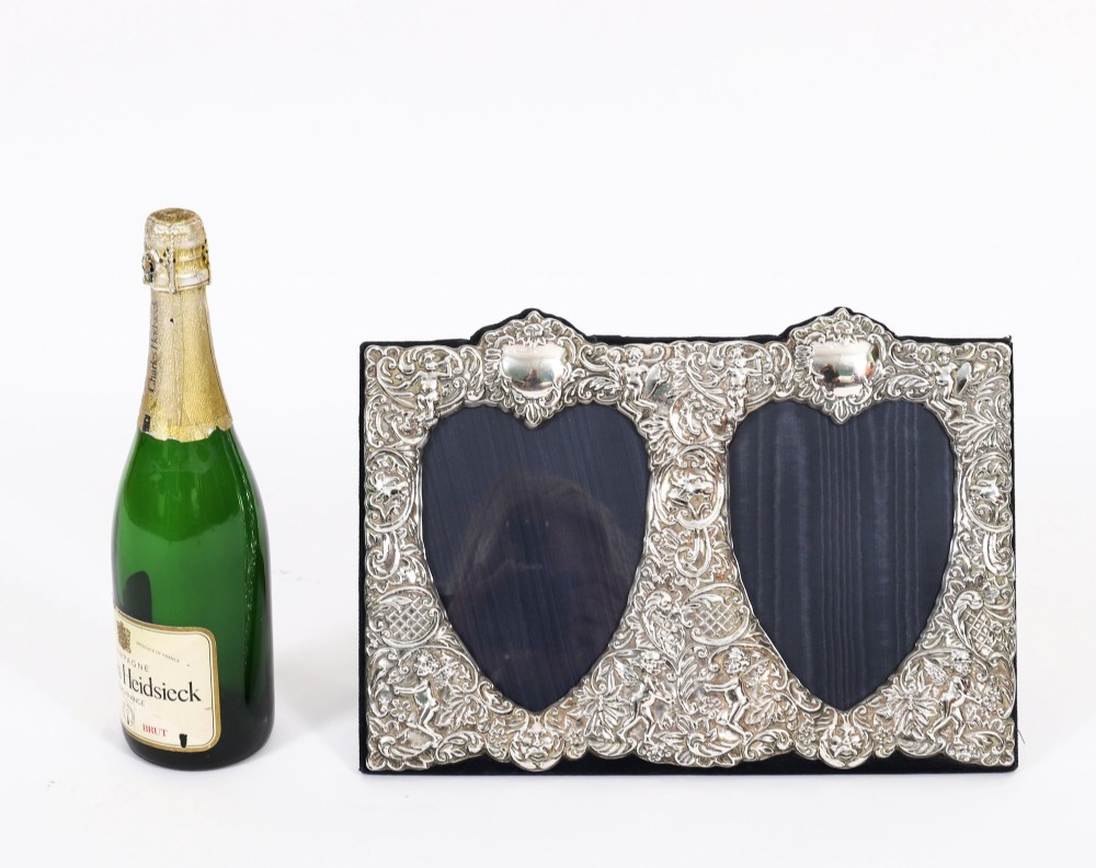 An antique sterling silver double aperture photo frame featuring heart-shaped openings, intricate repousse floral scrollwork, and cherub details mounted on dark velvet. View XIV