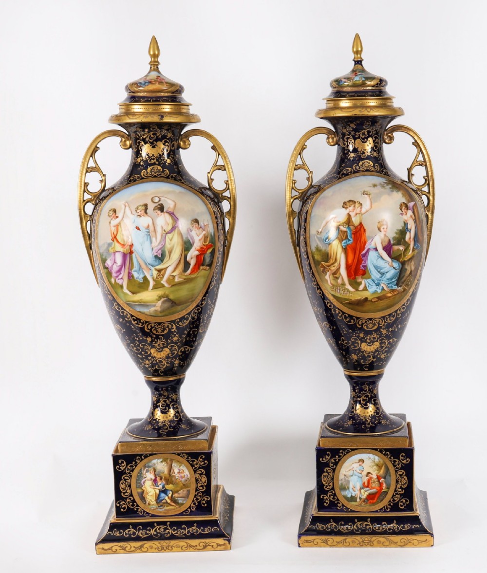 A pair of large 19th-century cobalt blue porcelain vases on matching square pedestals, featuring gilt scrollwork and hand-painted classical mythological scenes.