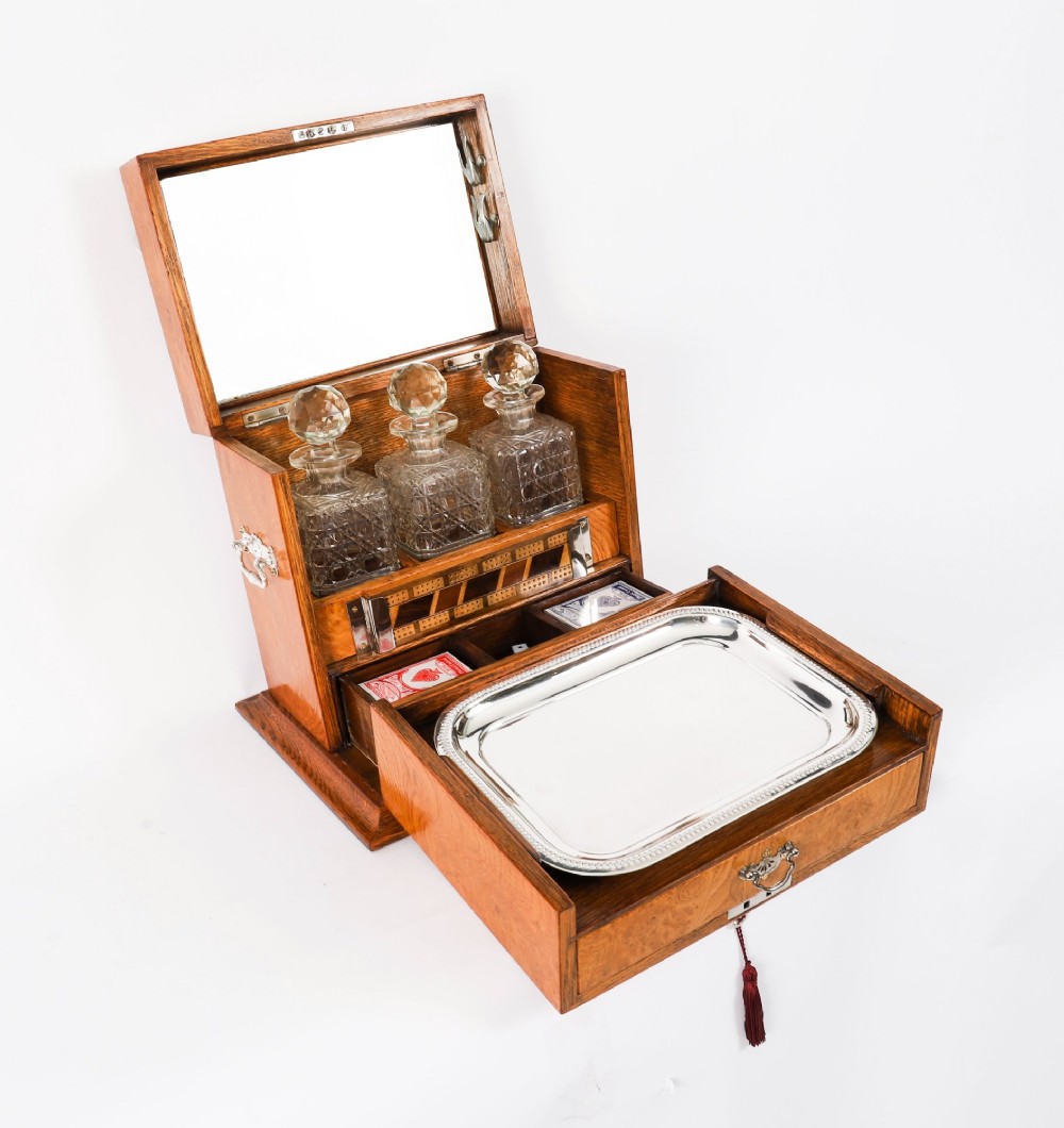 A 19th-century Victorian figured oak three-bottle tantalus open to reveal three cut glass decanters, a fitted cribbage board, and a drop-front drawer containing a silver-plated tray and playing cards.