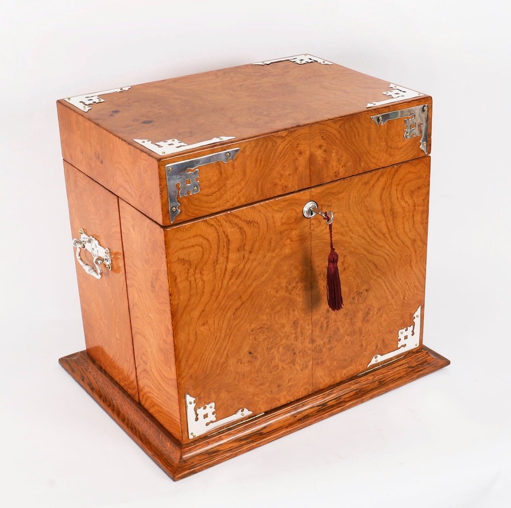 A 19th-century Victorian figured oak three-bottle tantalus open to reveal three cut glass decanters, a fitted cribbage board, and a drop-front drawer containing a silver-plated tray and playing cards. View IV