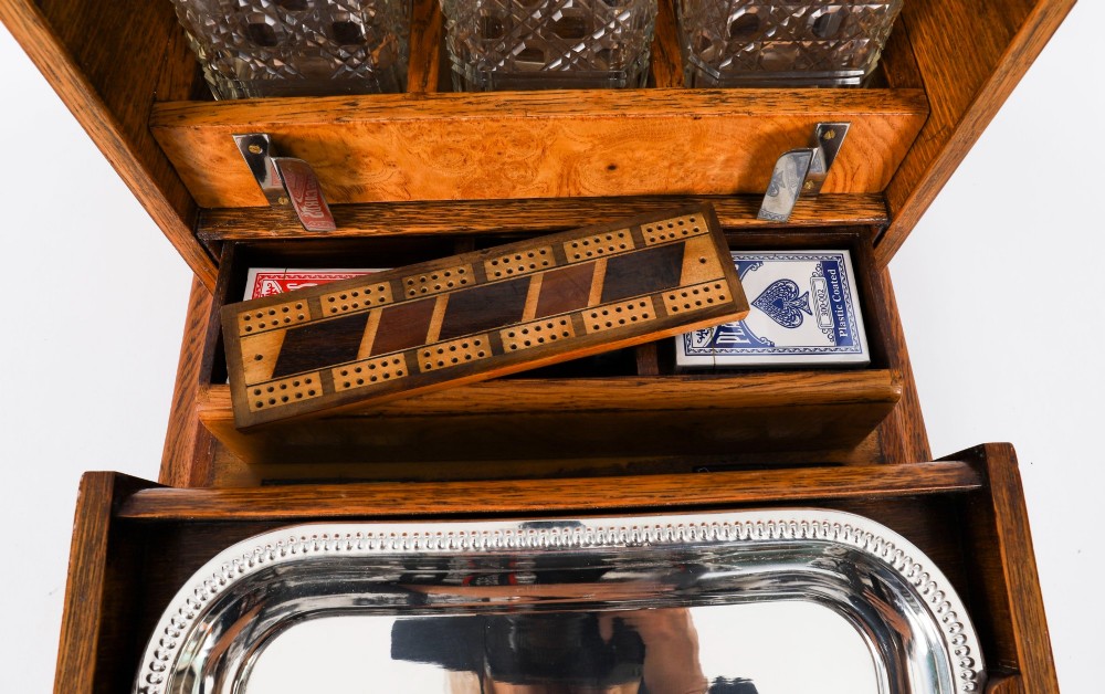 A 19th-century Victorian figured oak three-bottle tantalus open to reveal three cut glass decanters, a fitted cribbage board, and a drop-front drawer containing a silver-plated tray and playing cards. View V