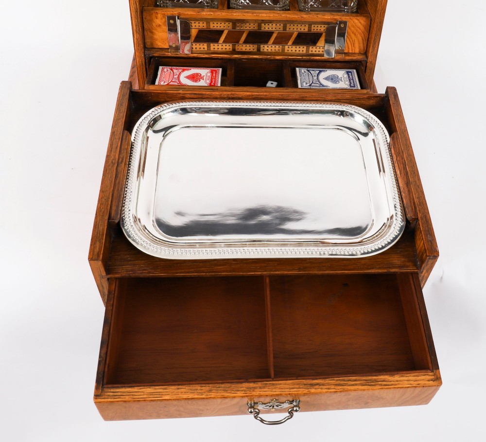 A 19th-century Victorian figured oak three-bottle tantalus open to reveal three cut glass decanters, a fitted cribbage board, and a drop-front drawer containing a silver-plated tray and playing cards. View VIII
