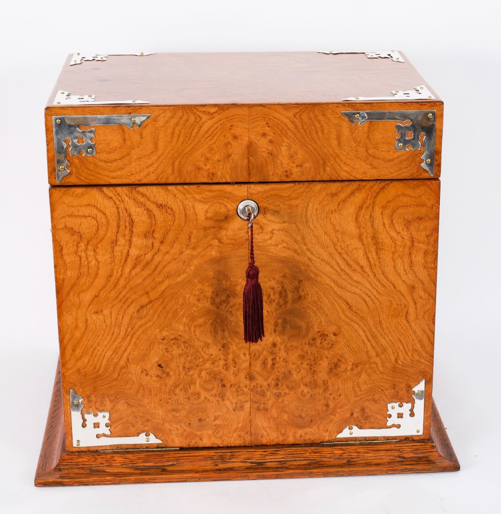 A 19th-century Victorian figured oak three-bottle tantalus open to reveal three cut glass decanters, a fitted cribbage board, and a drop-front drawer containing a silver-plated tray and playing cards. View IX