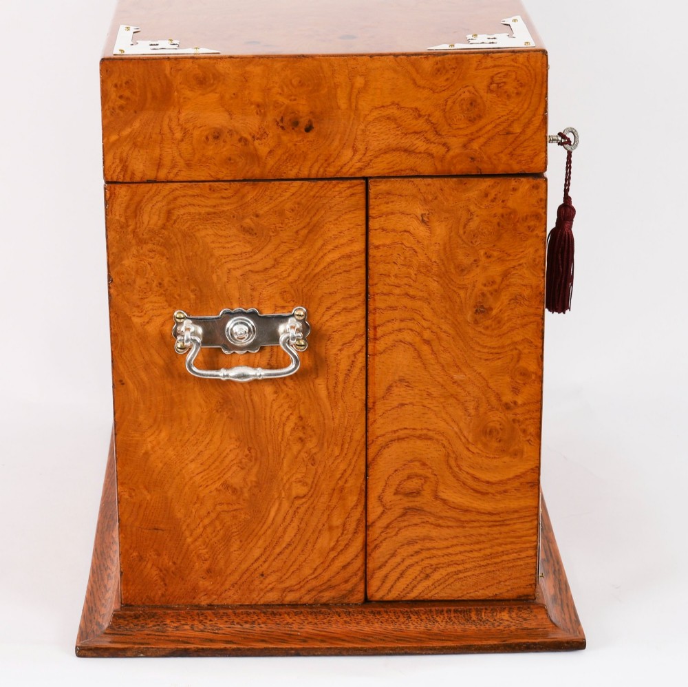 A 19th-century Victorian figured oak three-bottle tantalus open to reveal three cut glass decanters, a fitted cribbage board, and a drop-front drawer containing a silver-plated tray and playing cards. View XI