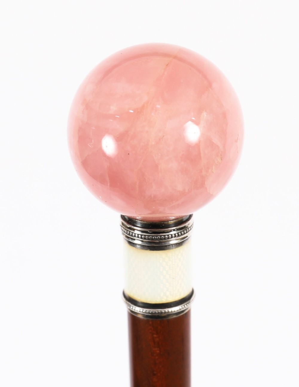 An antique walking stick featuring a spherical pink quartz pommel set above a white enamel collar with silver beaded borders and a dark wood shaft.