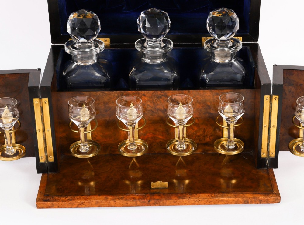 An open and closed 19th-century Victorian burr walnut tantalus box containing three cut crystal decanters, brass-mounted liqueur glasses, and a deep blue velvet lining. View IV