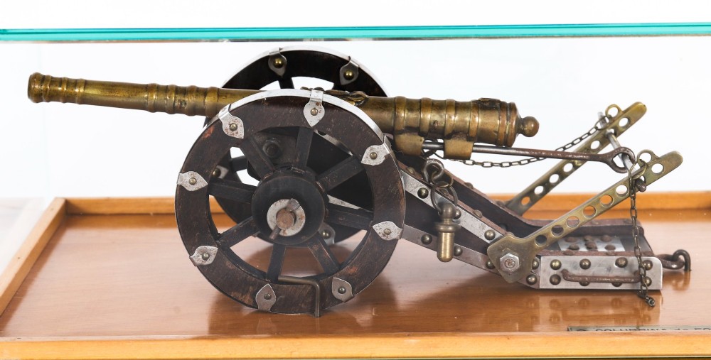A 19th-century bronze model cannon mounted on a dark wooden carriage and enclosed within a clear glass display case. View V