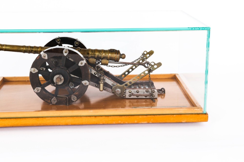 A 19th-century bronze model cannon mounted on a dark wooden carriage and enclosed within a clear glass display case. View VI