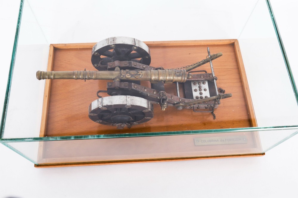 A 19th-century bronze model cannon mounted on a dark wooden carriage and enclosed within a clear glass display case. View IX