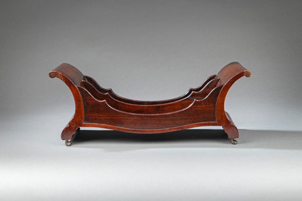 A Georgian mahogany cheese coaster featuring a curved boat-shaped form with internal dividers, scrolled ends, and small brass castors on the base.