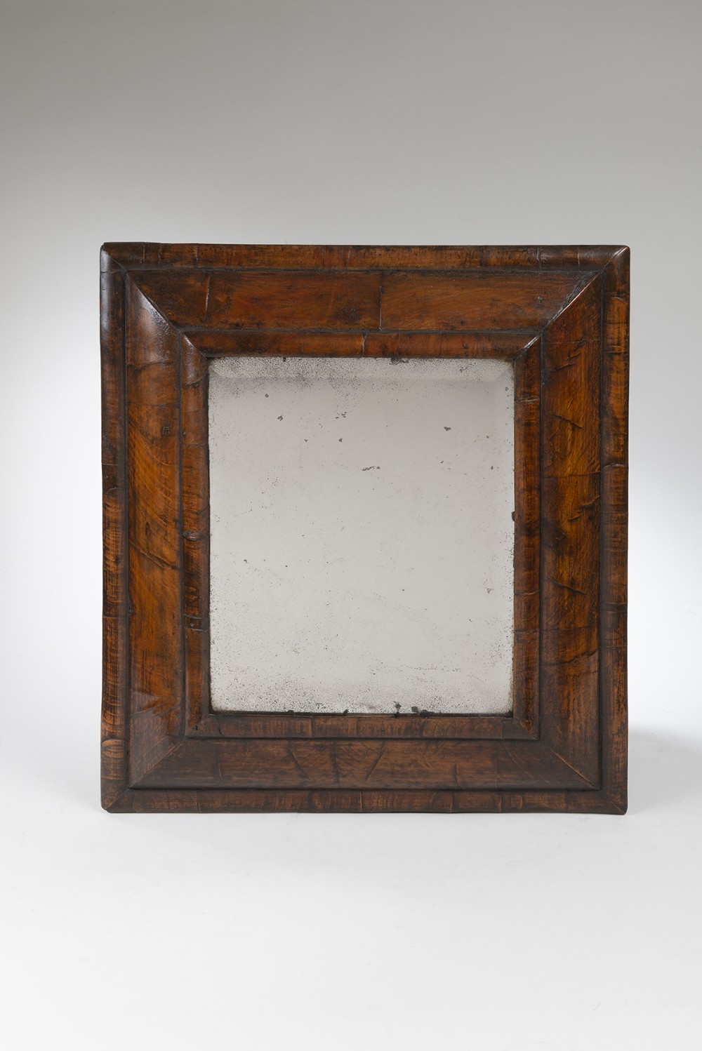 A rectangular wall mirror featuring a pronounced, sloped walnut-veneered cushion frame surrounding an aged glass plate.