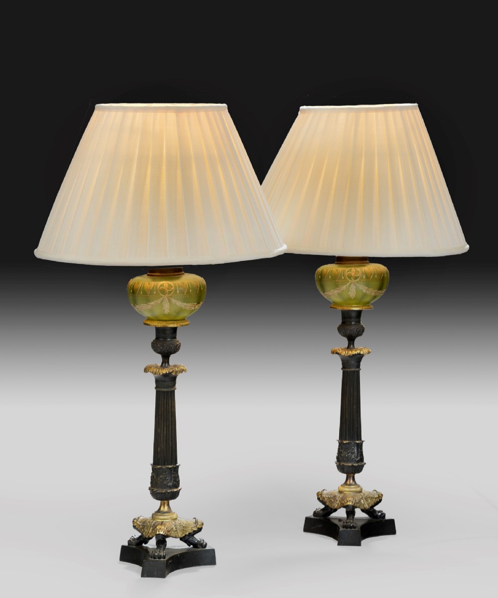 A pair of antique Regency style table lamps featuring fluted dark bronze columns, gilt-ormolu mounts, green decorated glass elements, and cream pleated shades.