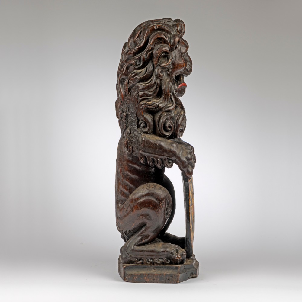 A seventeenth-century dark carved oak lion newel post finial seated upright while holding a painted heraldic shield with yellow, red, and black coat of arms details. View II