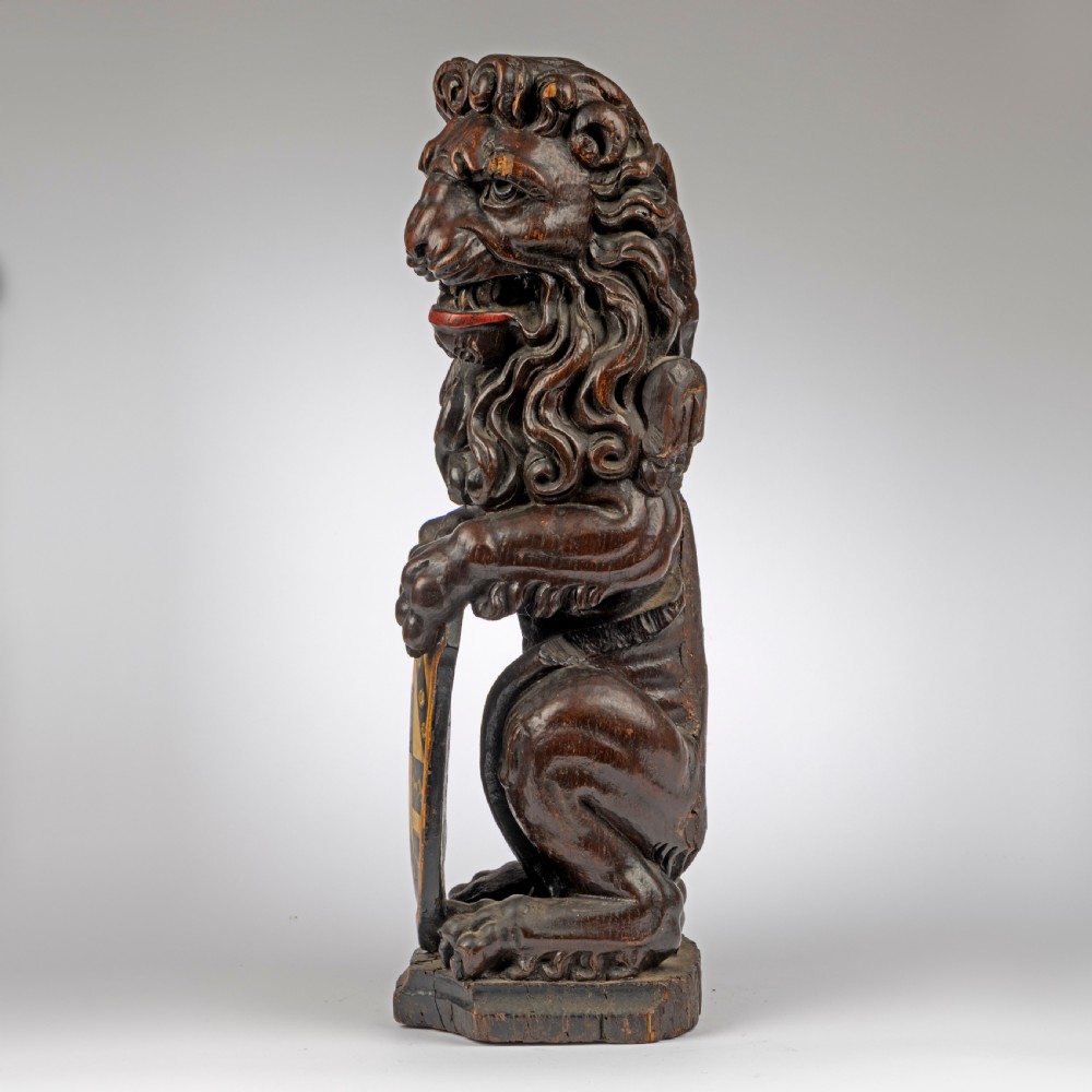 A seventeenth-century dark carved oak lion newel post finial seated upright while holding a painted heraldic shield with yellow, red, and black coat of arms details. View IV